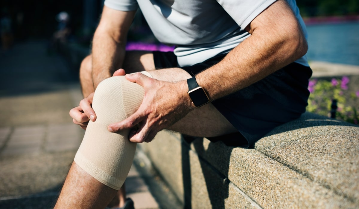 Picture of a man with a knee wrap, sitting down outside on steps holding his knee