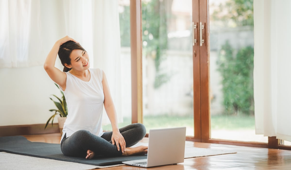 image of a woman sitting on a yoga mat, stretching while sitting in front of a laptop